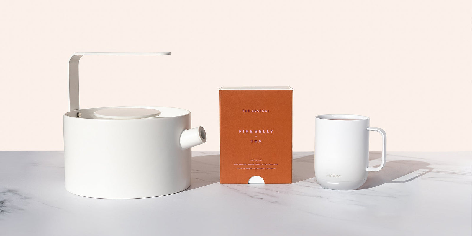 A white geometric tea kettle sits on the left, a red Firebelly Tea Sampler box sits in the middle, and an Ember Mug² in white sits on the right on a grey and cream background.